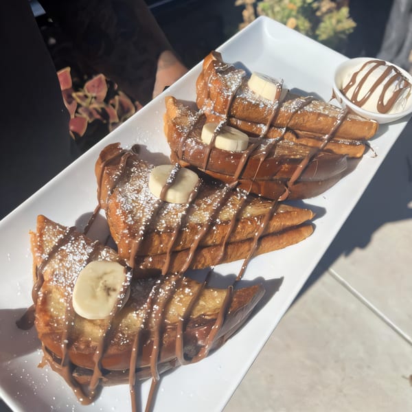 French toast with chocolate drizzle, powdered sugar, banana slices, and a side of whipped cream.